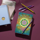 Sri Yantra Notebook with vibrant sacred geometry cover placed on a purple desk with pens and other notebooks, ideal for journaling and gifting.